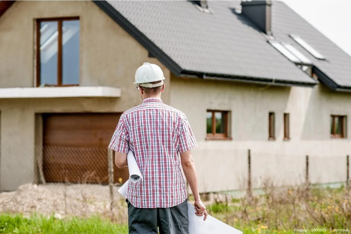 Mann mit Helm auf dem Kopf steht vor einem Haus - In diesem Beitrag erfahren Sie wissenswerte Infos darüber, was Sie beim Kellerbau beachten sollten und hilfreiche Tipps und Tricks für die Kellerplanung.